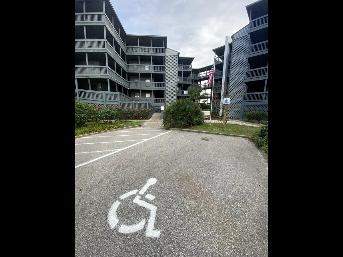 The image highlights a building entrance with visible handicap parking, accentuated landscaping, and accessible pathways. The mood is welcoming and inclusive, suggesting ease of access and a sense of community. The well-kept surroundings reinforce the property’s commitment to a clean, comfortable stay.