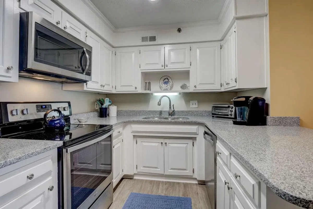 This image showcases a bright, welcoming kitchen with stainless steel appliances, granite countertops, white cabinetry, a kitchen sink, and a cozy blue rug. The open layout is ideal for preparing meals or socializing, and it reflects a space where cooking, gathering, and casual dining blend together seamlessly.