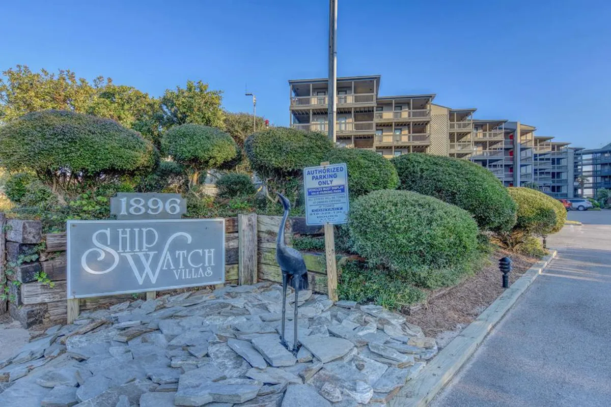 The entrance to Ship Watch Villas is framed by lush landscaping and a decorative bird sculpture, with the community sign welcoming guests. The scene is tranquil, creating a strong sense of arrival and hinting at both the peaceful setting and the opportunity to gather in common outdoor spaces.