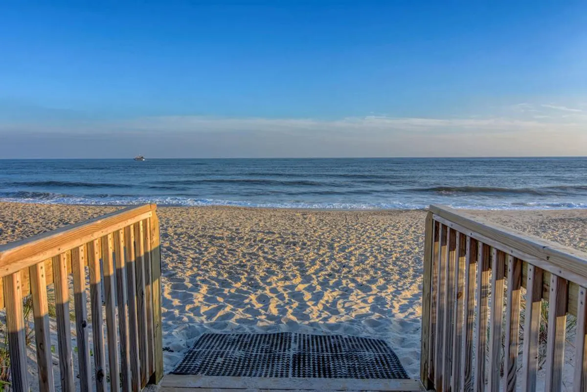This image shows a wooden walkway leading directly onto a sandy beach with gentle ocean waves and a clear sky in the background. The view highlights the seamless transition from property to beach, emphasizing accessibility and the natural beauty of North Topsail Beach.