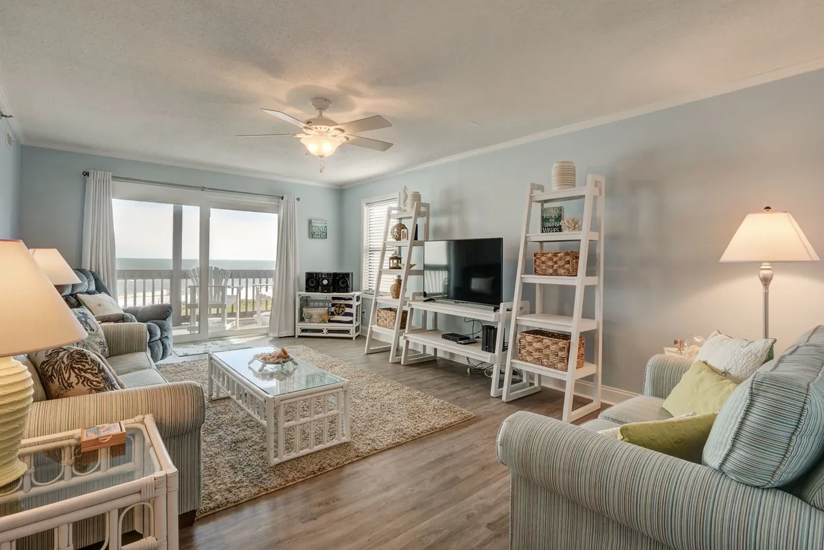 A bright and airy living space is showcased here, with large windows letting in plenty of natural light, a cozy seating area, decorative shelving, and a glass coffee table. The decor is subtle and beach-inspired, contributing to the tranquil mood.