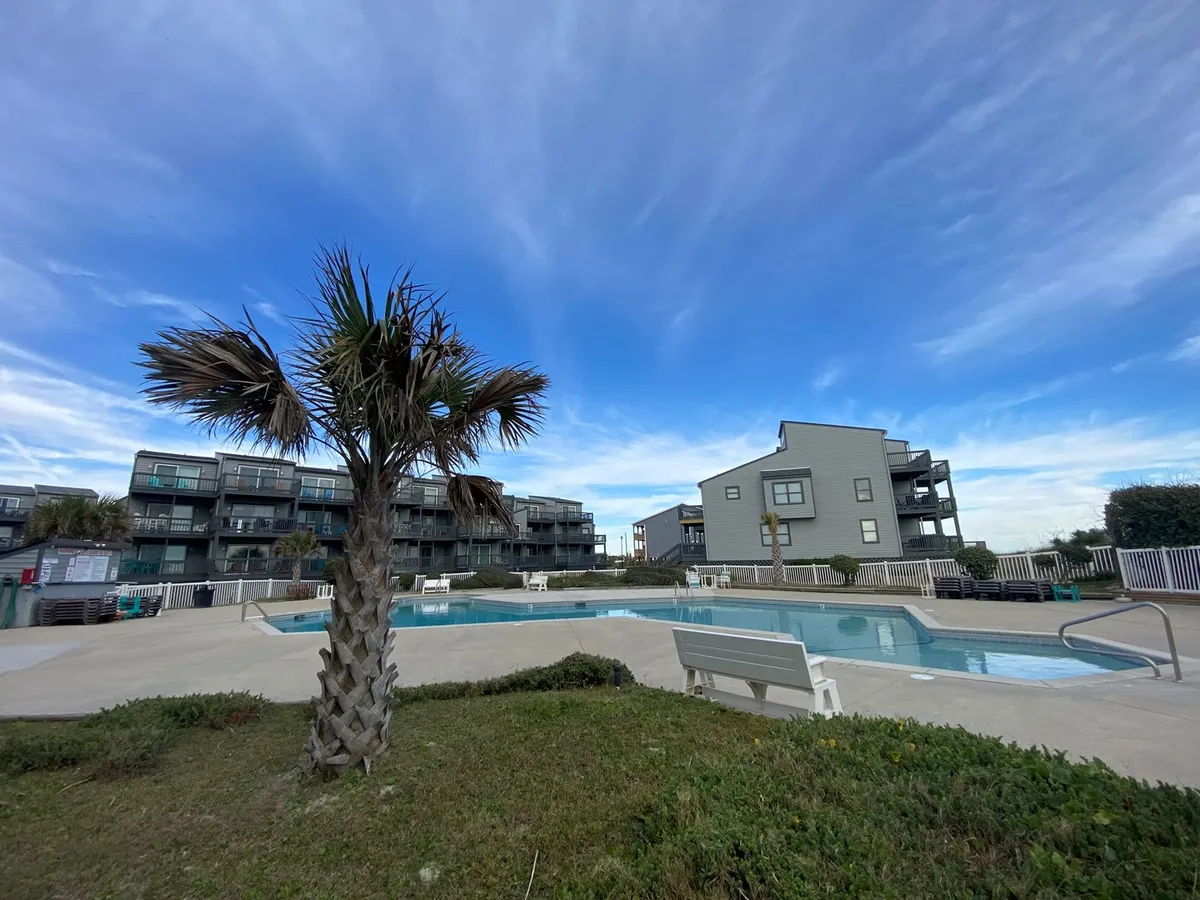 A sunlit swimming pool area surrounded by palm trees and modern buildings, inviting guests to relax, socialize, or watch a sunset by the water after a day exploring the island.