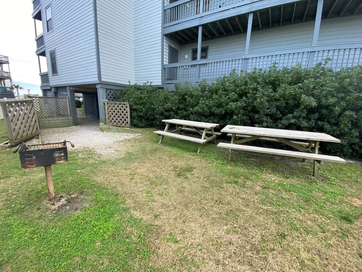 An outdoor grilling area features picnic tables and a communal grill, bordered by greenery and the condo buildings. The setup encourages al fresco dining and socializing in a relaxed, communal setting.