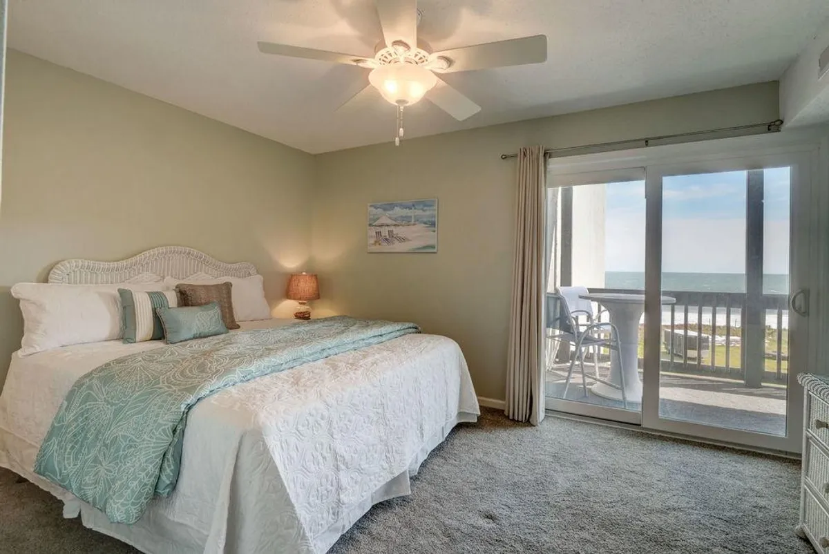 A spacious bedroom with a large bed, decorative pillows, and a ceiling fan, featuring sliding glass doors that lead out to the balcony. The view hints at the ocean beyond, and the room feels bright, airy, and peaceful.
