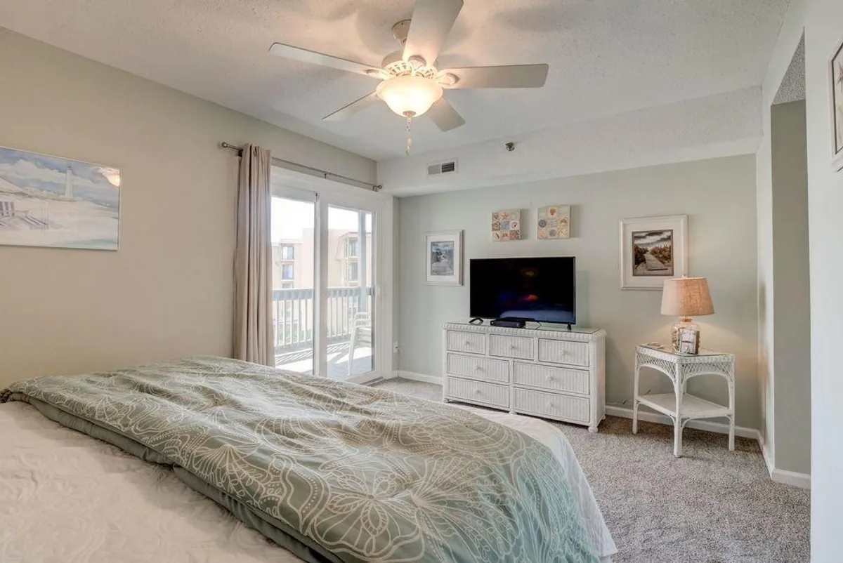 An inviting bedroom featuring a bed with a patterned duvet and decorative pillows, a television unit, and sliding glass doors that open directly onto the balcony. Bright natural light pours in, connecting the indoor sanctuary to the oceanfront retreat outside.