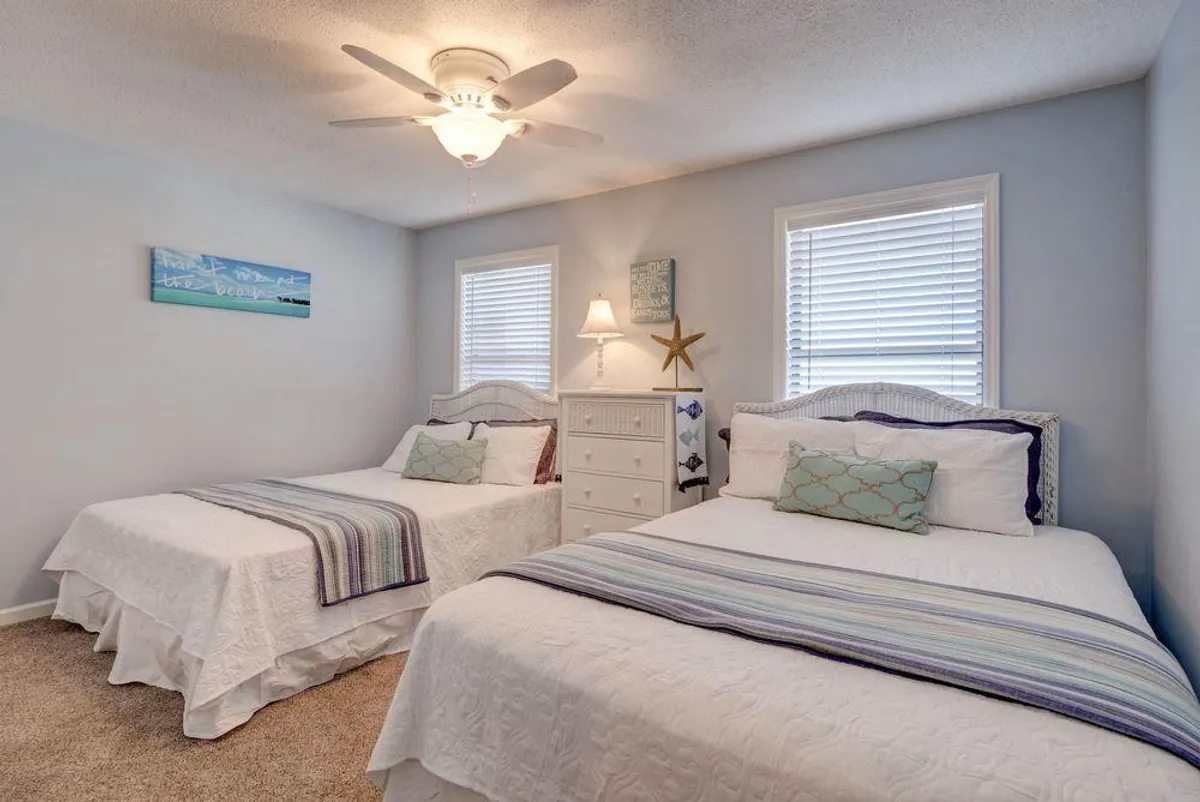 A welcoming guest bedroom with two twin beds dressed in crisp white linens, accented by decorative pillows. A nightstand with a lamp sits between the beds, and whimsical, coastal-themed wall art and a starfish decoration reinforce the beach atmosphere. Soft natural light fills the room, creating a comfortable setting for families or friends.